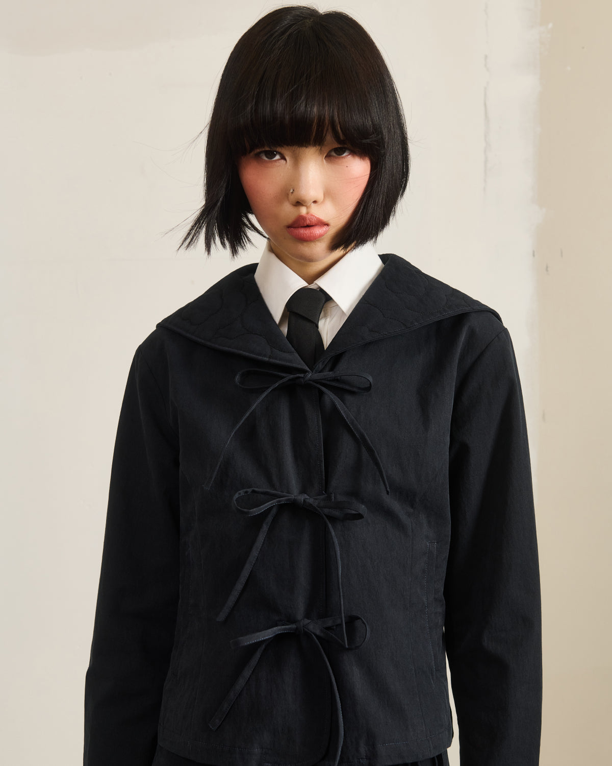 A person with short black hair and blunt bangs wears the Sundae School BLACK SAILOR COLLAR UNIFORM JACKET, featuring three bow ties and a wide collar over a white shirt and black tie, standing by a light-colored textured wall.