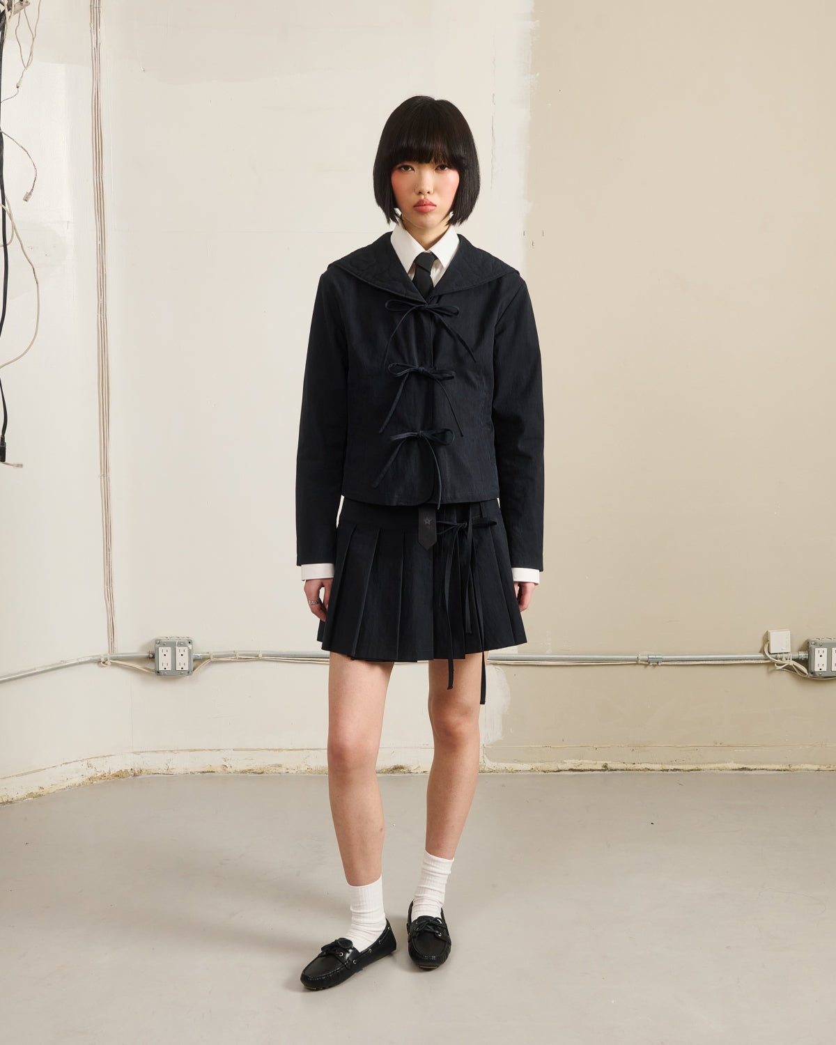 A person with a short black bob wears Sundae School's BLACK UNIFORM APRON SKIRT over a dark sailor-style jacket, white shirt, and black tie, standing in a plain room with exposed wires and outlet panels.
