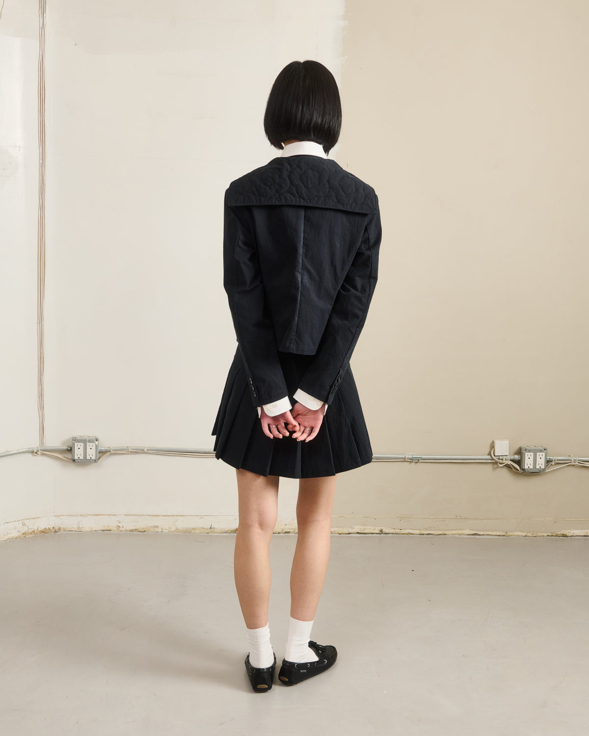 A person with short black hair faces a beige wall, wearing Sundae School’s BLACK UNIFORM APRON SKIRT with a matching jacket, white socks, and black shoes. Their arms are relaxed at their sides in a sparse room with visible outlets and cables.
