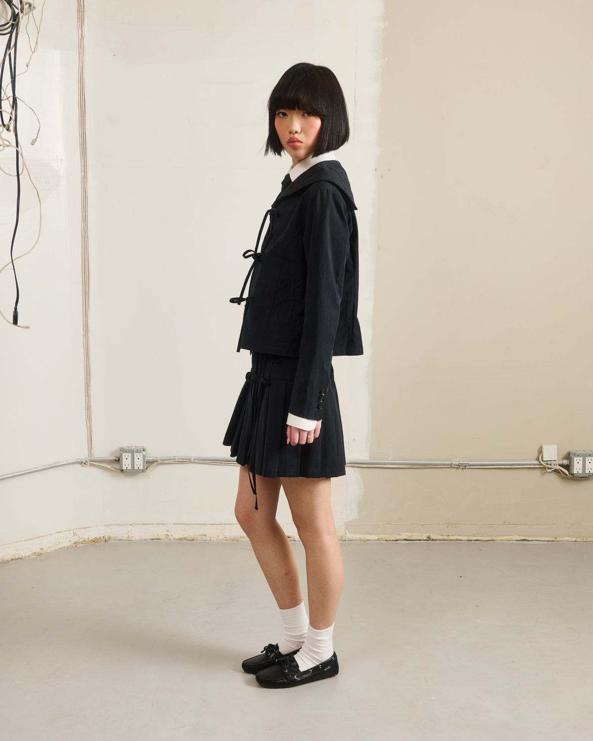 A young woman with a short black bob stands in a minimal room, wearing Sundae School’s BLACK UNIFORM APRON SKIRT with a black jacket, white socks, and black shoes. She faces slightly to the side; exposed wires are visible on the wall behind her.