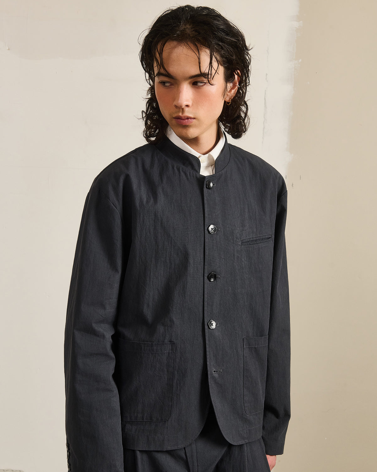 A person with wavy dark hair wears the CHARCOAL STAND COLLAR UNIFORM JACKET by Sundae School over a white shirt, standing against a light beige background and looking to the side with a neutral expression.