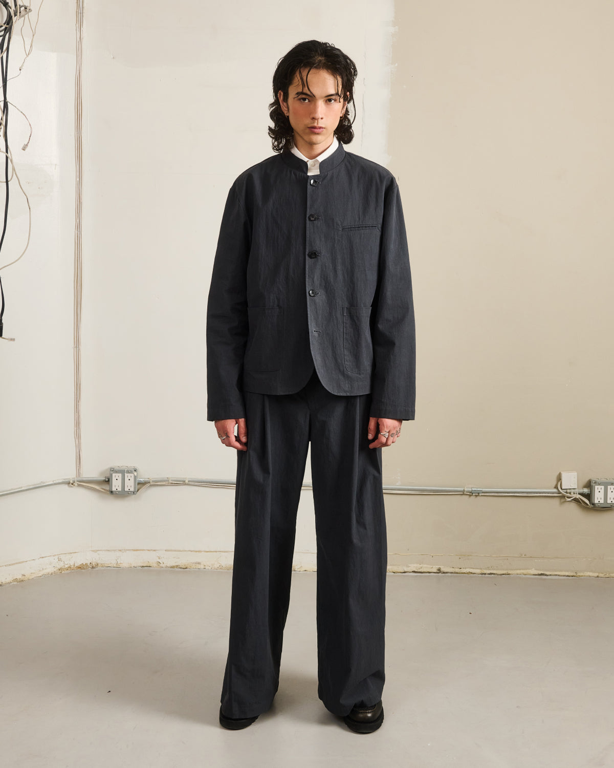 A young person with long dark hair faces forward, wearing Sundae School CHARCOAL PLEATED UNIFORM SLACKS inspired by Korean school uniforms and a dark grey mandarin collar jacket.