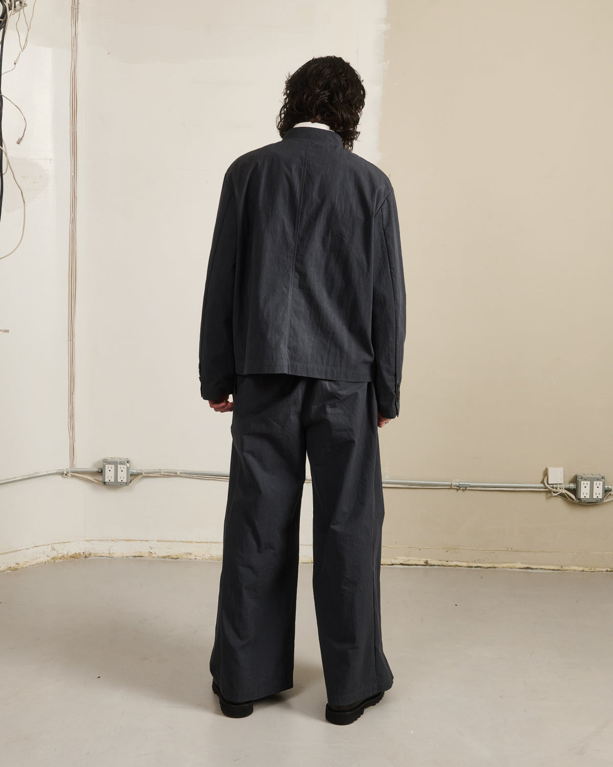 A person with curly hair faces a beige wall, wearing a loose-fitting dark gray jacket and Sundae School’s CHARCOAL PLEATED UNIFORM SLACKS. Electrical outlets and exposed wiring are visible along the lower part of the wall.