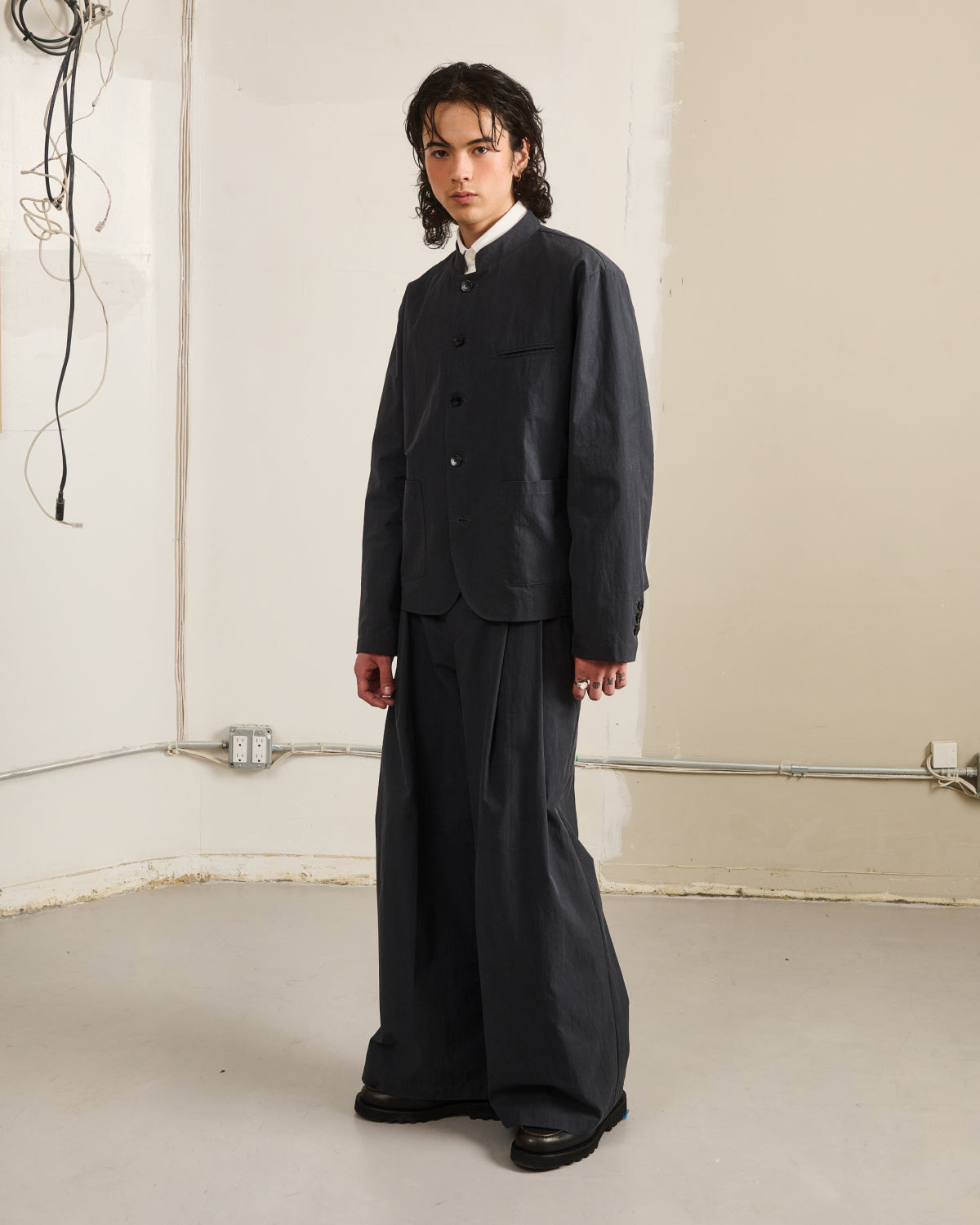 A person with long dark hair stands indoors against a plain wall, wearing Sundae School's CHARCOAL STAND COLLAR UNIFORM JACKET and wide-leg trousers in a cotton-nylon blend, over a white shirt. Exposed wires and outlets are visible on the walls.