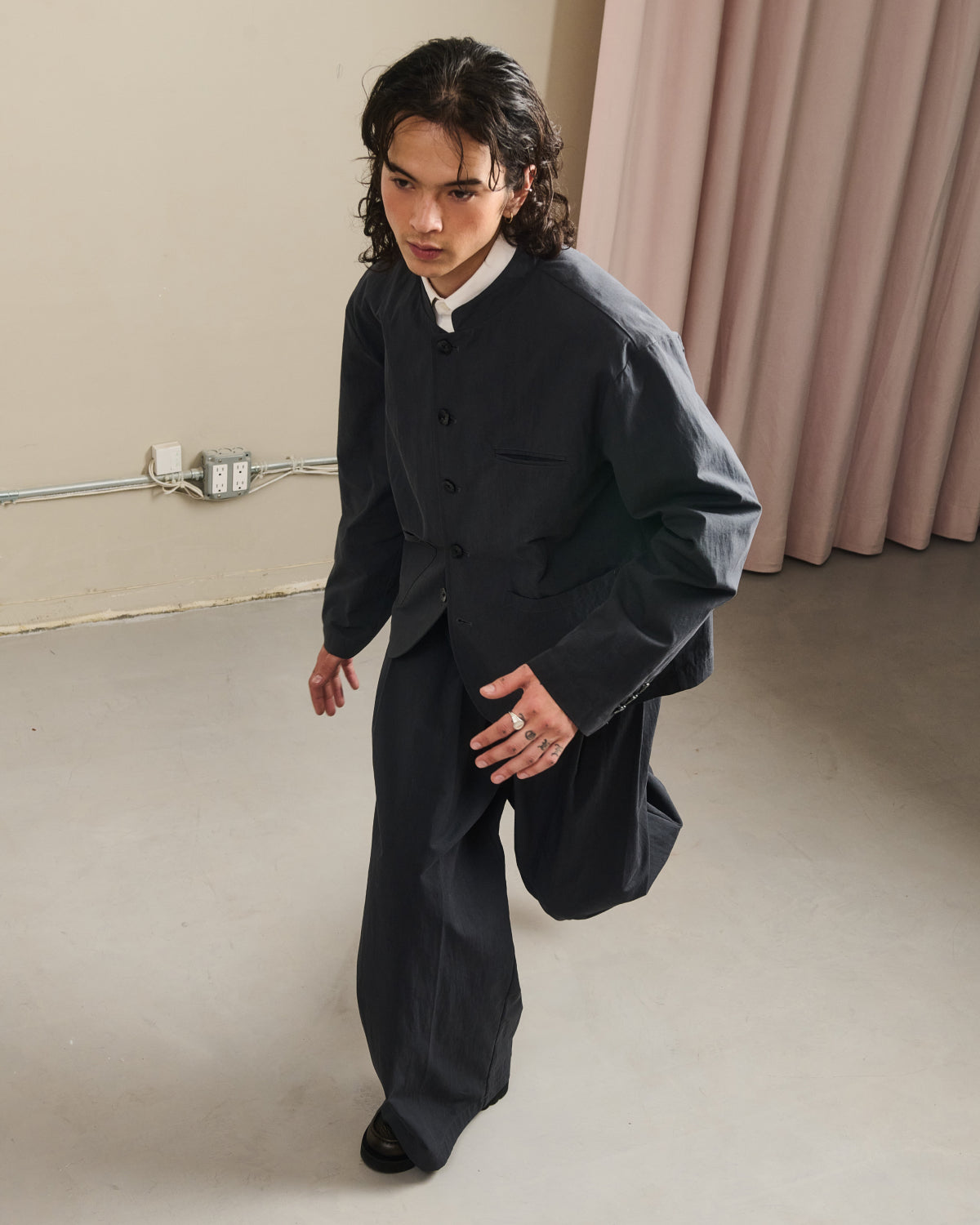 A person with long dark hair wears Sundae School’s CHARCOAL PLEATED UNIFORM SLACKS and a white shirt, posing indoors on a light gray floor near pink curtains and a beige wall with visible outlets and cords.