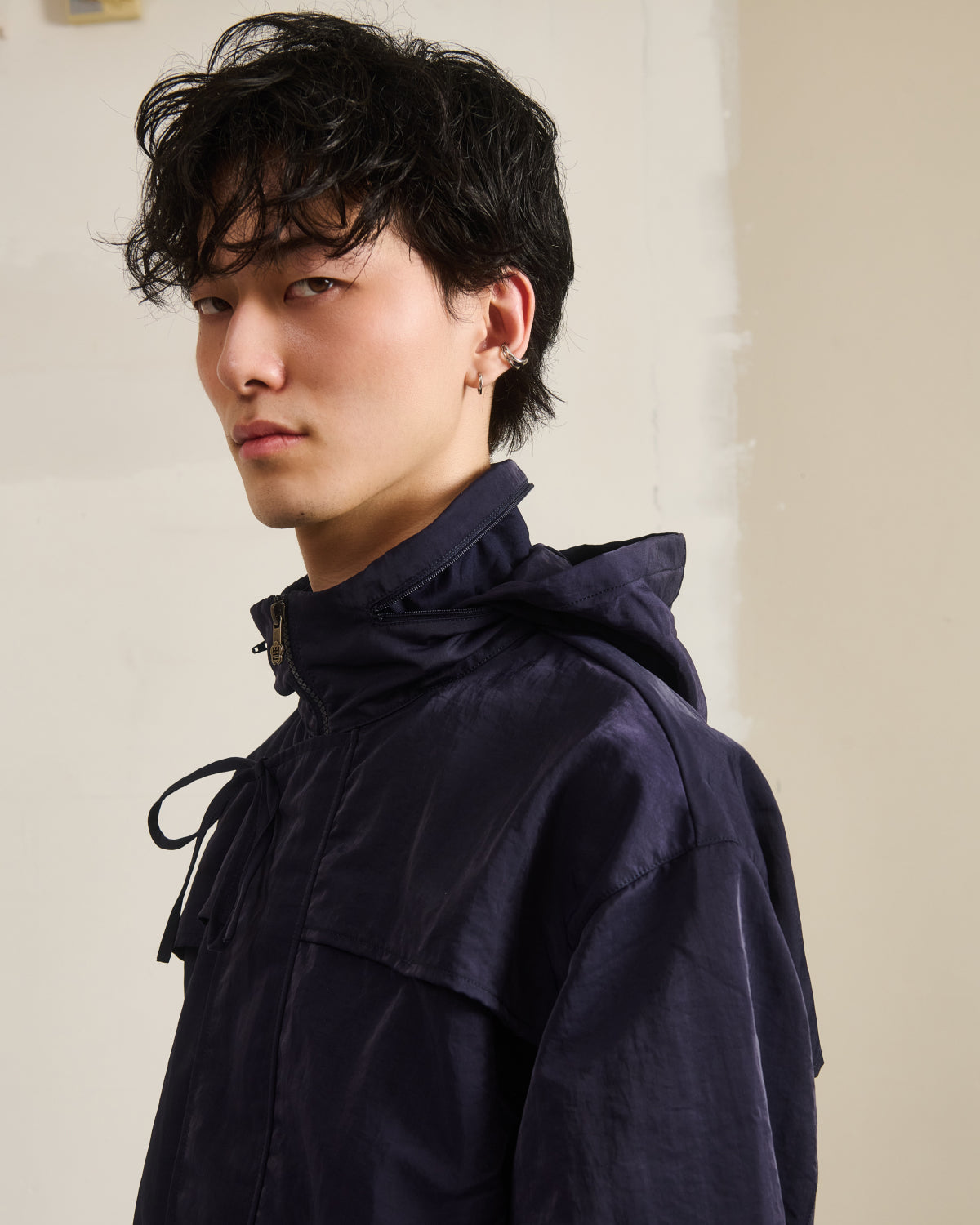 A young man with black, tousled hair and an earring wears the NAVY HANBOK TRACK JACKET by Sundae School, featuring a custom 떨 zipper pull, as he stands against a light wall and looks neutrally over his shoulder at the camera.