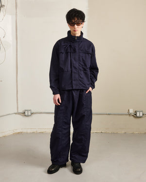 A person stands indoors against a beige wall, hands in pockets, wearing Sundae School NAVY HANBOK BALLOON TRACK PANTS with adjustable hems and a loose-fitting navy jacket. Electrical outlets and cords are visible in the background.