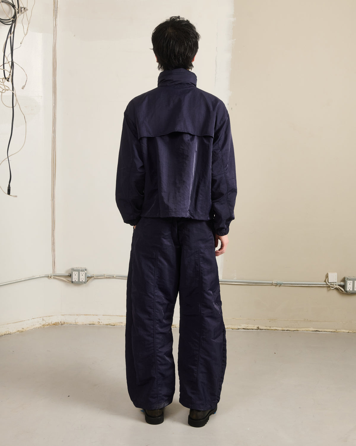 A person with short dark hair faces a beige wall, wearing Sundae School’s NAVY HANBOK TRACK JACKET and matching wide-leg pants. Exposed wires and electrical outlets are visible on the left side of the wall.