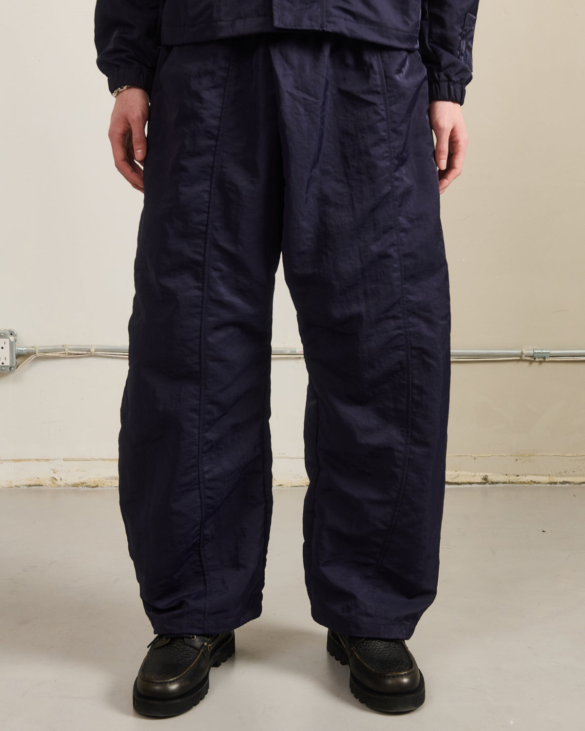 A person stands indoors wearing Sundae School's NAVY HANBOK BALLOON TRACK PANTS and black shoes. The lower body is shown against a plain background with visible baseboard and pipes; the photo is cropped at the shoulders.