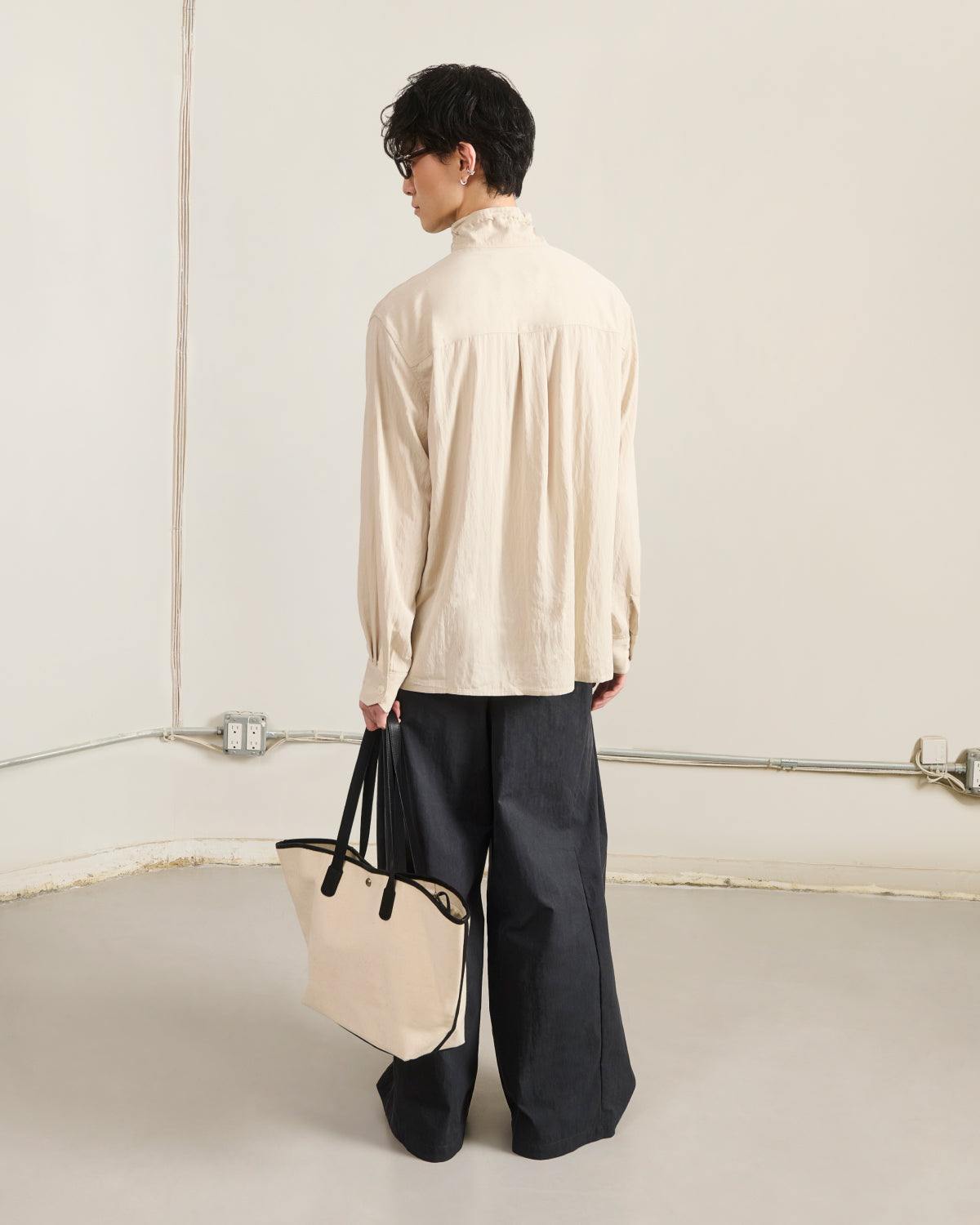 A person with short dark hair wears the Sundae School IVORY HIGH NECK TIE SHIRT and wide-leg dark pants, standing in a minimalist room while holding a beige tote bag with black handles.