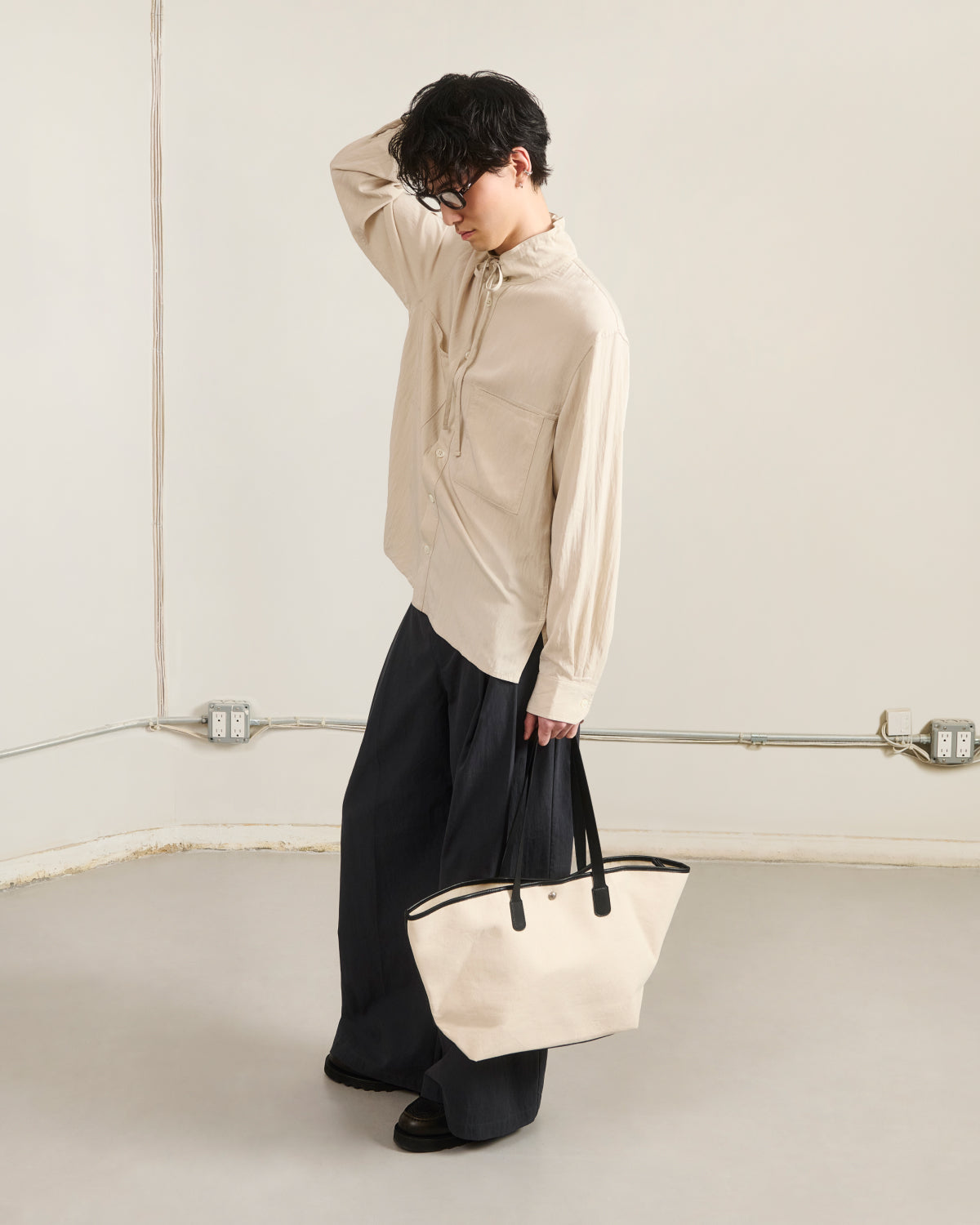 A person with short dark hair and glasses stands indoors, wearing the Sundae School IVORY HIGH NECK TIE SHIRT, wide-legged dark pants, and black shoes, holding a light tote bag with one hand touching their head.