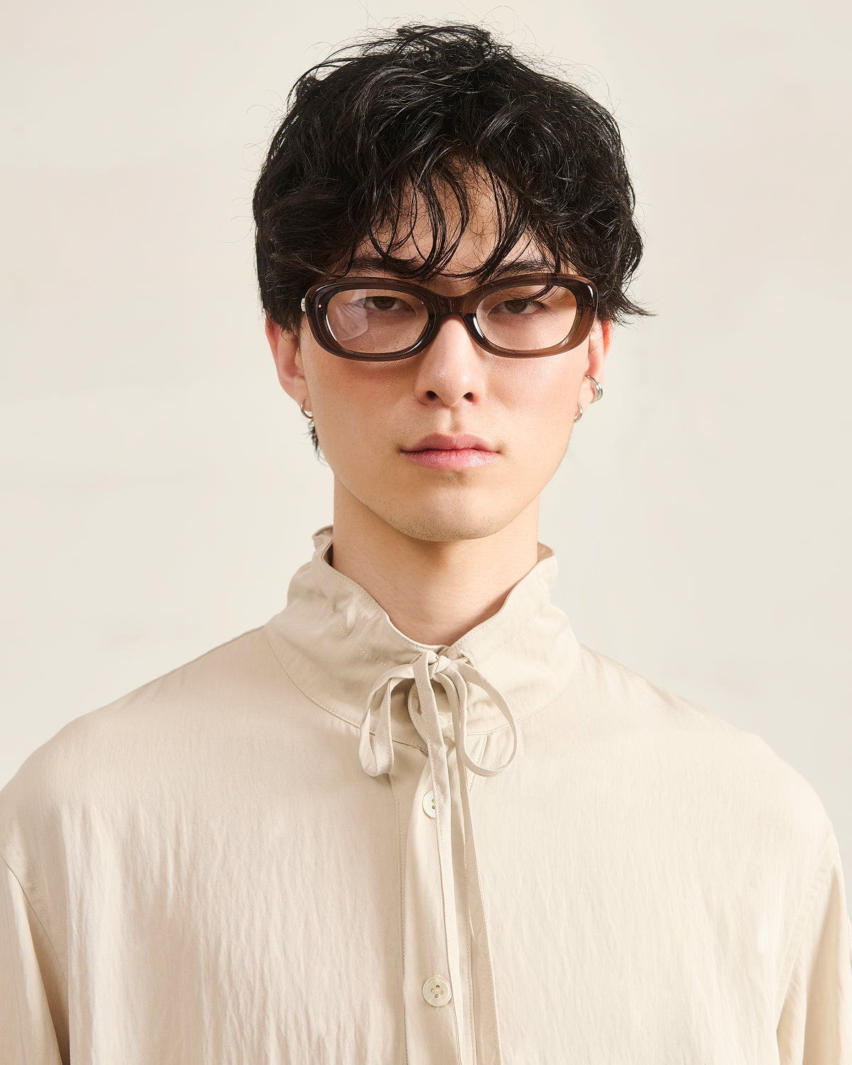 A person with wavy dark hair and glasses models the Sundae School IVORY HIGH NECK TIE SHIRT, a light beige convertible top with a neck tie detail and summer-weight fabric, posed against a plain light background.