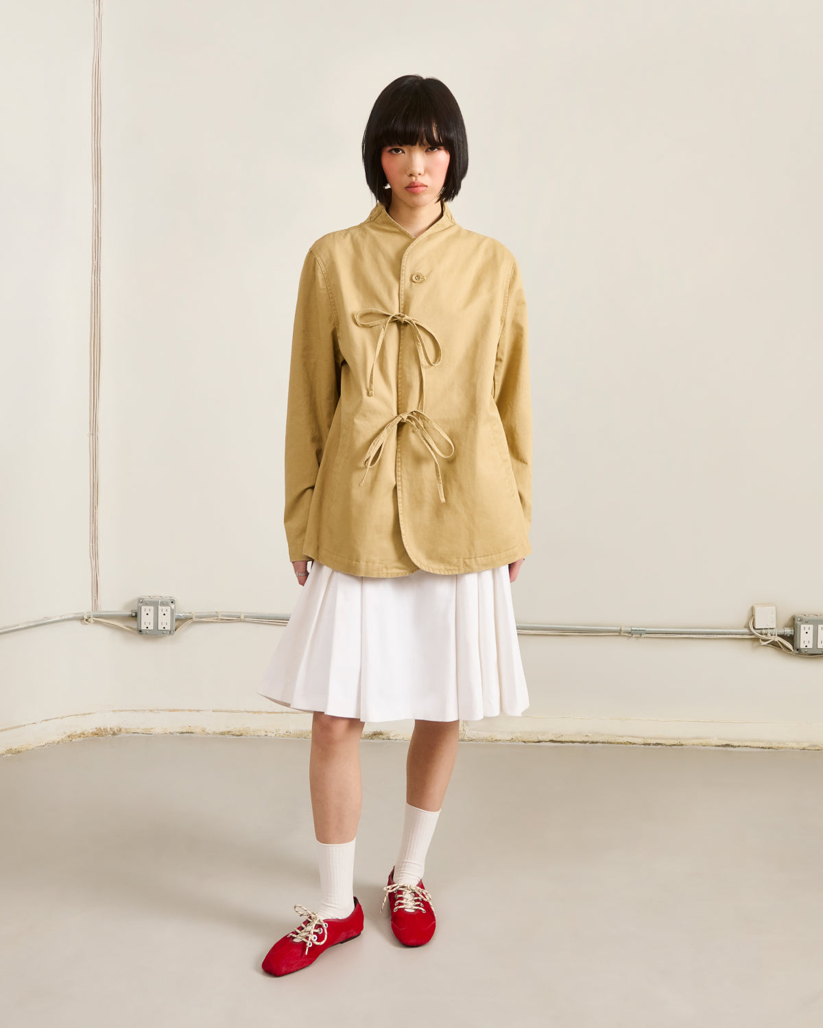 A person with a short black bob wears Sundae School’s TAN JEOGORI TIE JACKET, paired with a white pleated skirt, white socks, and red shoes, standing against a cream wall with exposed outlets and conduit.