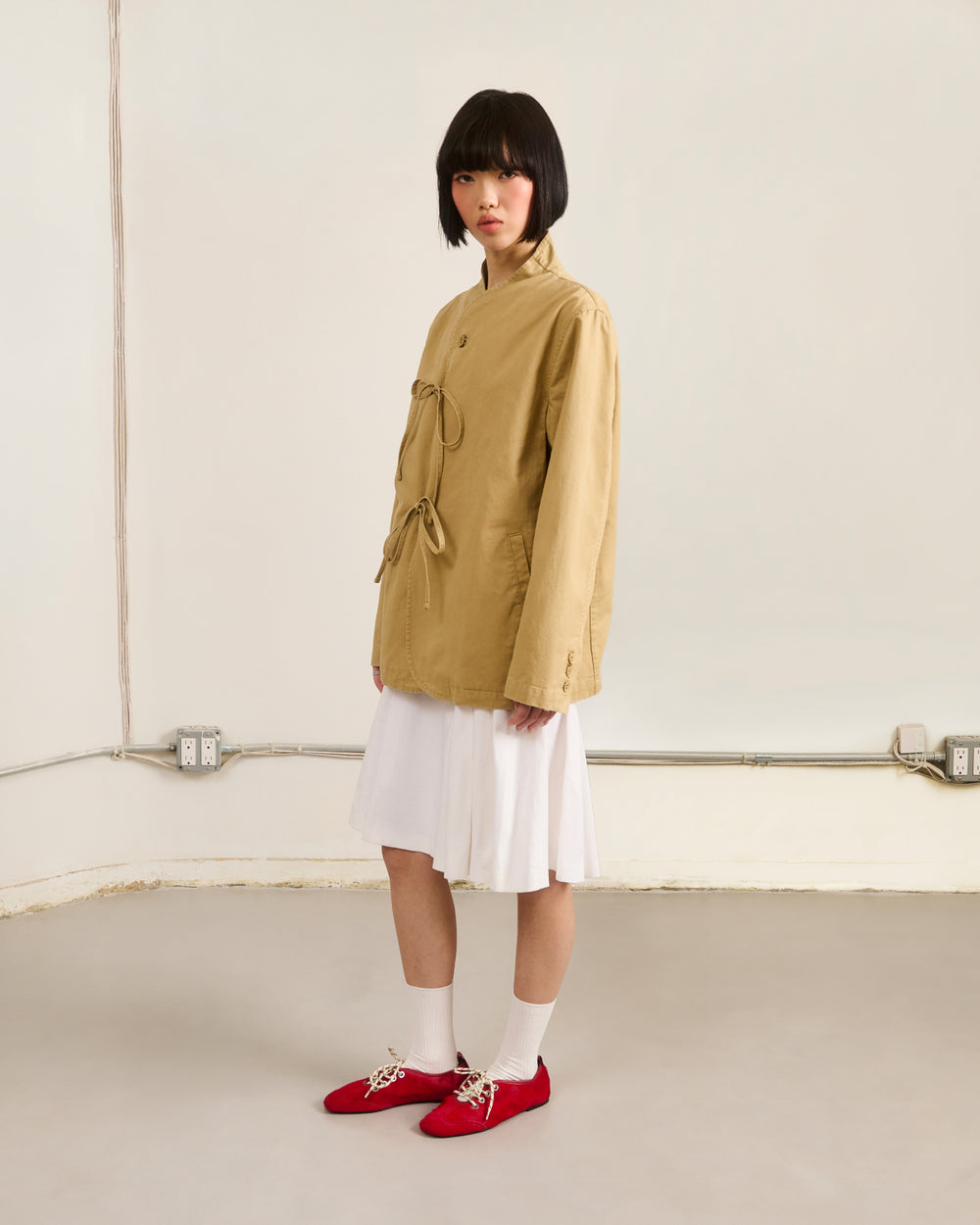 A person with a blunt bob stands indoors in a Sundae School TAN JEOGORI TIE JACKET over a white knee-length dress, paired with white socks and bright red shoes, against a plain backdrop featuring visible electrical outlets and cables.