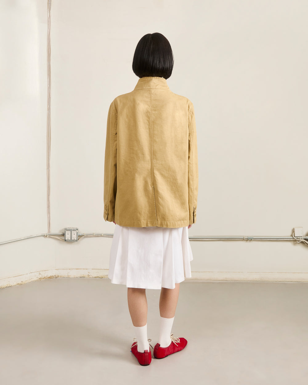 A person with short black hair wears the Sundae School TAN JEOGORI TIE JACKET, paired with a white knee-length skirt, white socks, and red shoes, standing by a beige wall with visible outlets and exposed cables.
