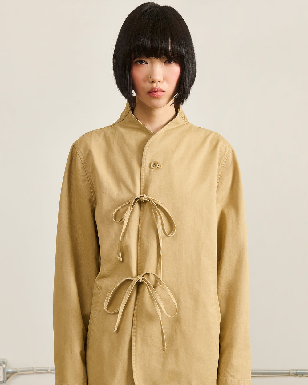 A person with a short black bob wears the TAN JEOGORI TIE JACKET by Sundae School, featuring a monk collar and two front ties for a modern jeogori look, standing against a plain light background.