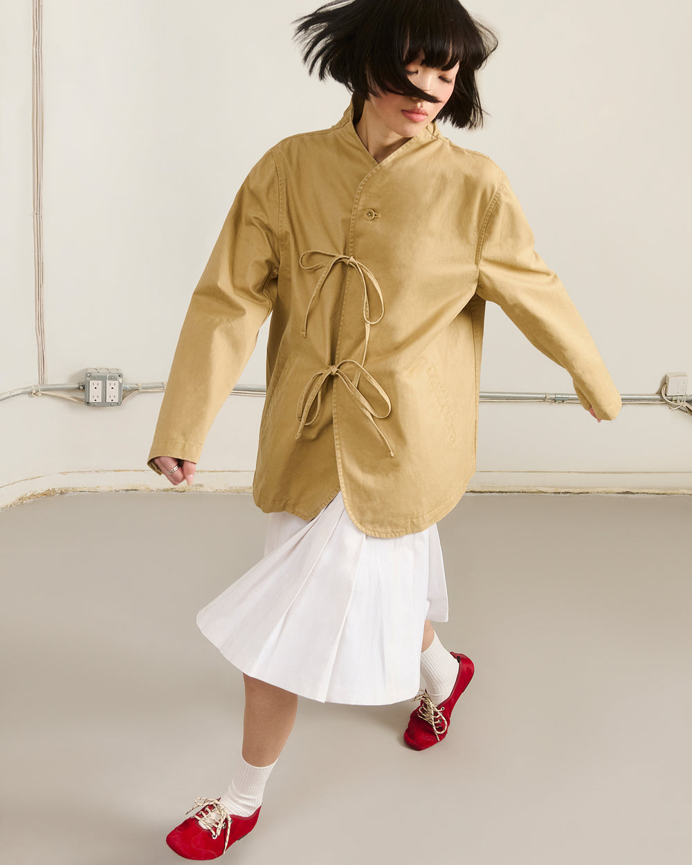 A person with short dark hair wears the Sundae School TAN JEOGORI TIE JACKET, styled over a white pleated skirt with white socks and red shoes. They are mid-step in a minimalist room with light-colored walls and floor.