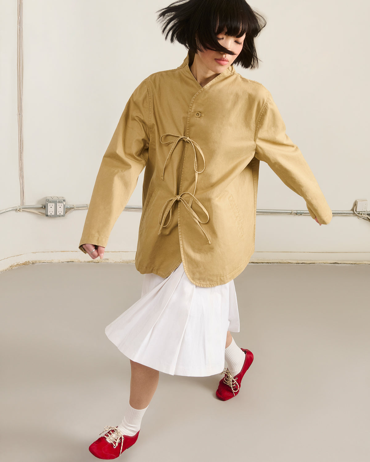 A person with short dark hair wears the Sundae School TAN JEOGORI TIE JACKET, styled over a white pleated skirt with white socks and red shoes. They are mid-step in a minimalist room with light-colored walls and floor.