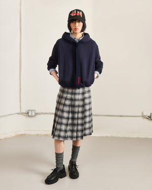 Indoors, a person wears the Sundae School NAVY KNIT TIE HOODIE with a black bucket hat, checkered knee-length skirt, gray socks, and black shoes. Hands in pockets; outlets and pipes are visible on the plain wall behind them.