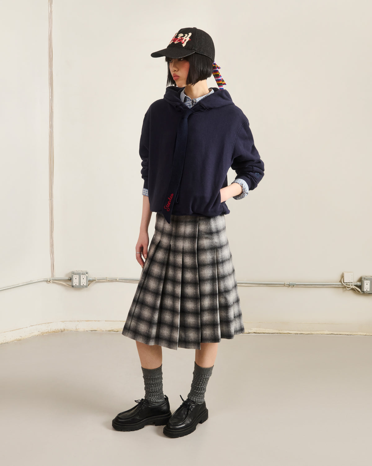 Wearing the Sundae School NAVY KNIT TIE HOODIE over a collared shirt, a black and white plaid pleated skirt, gray socks, black shoes, and a black baseball cap, a person stands in a minimalist room looking to the side.