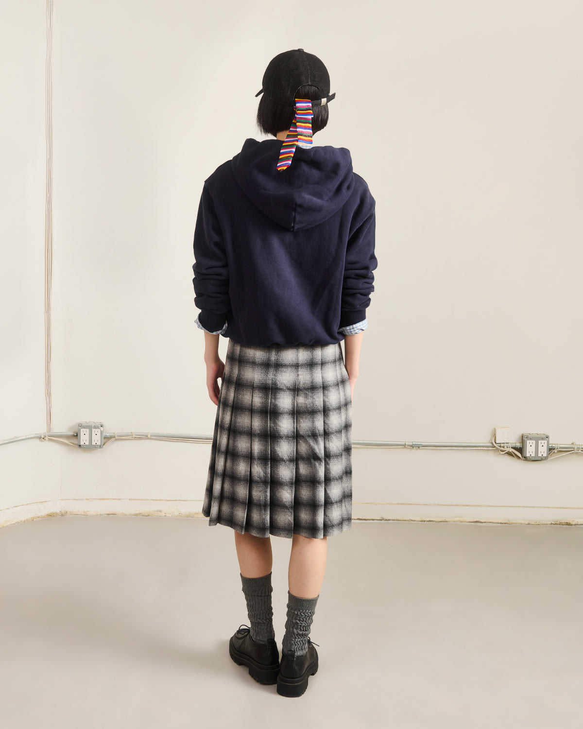A person stands facing away in a plain room with exposed conduits, wearing the Sundae School NAVY KNIT TIE HOODIE, a black cap with striped ribbon, black-and-white plaid skirt, gray socks, and black shoes.
