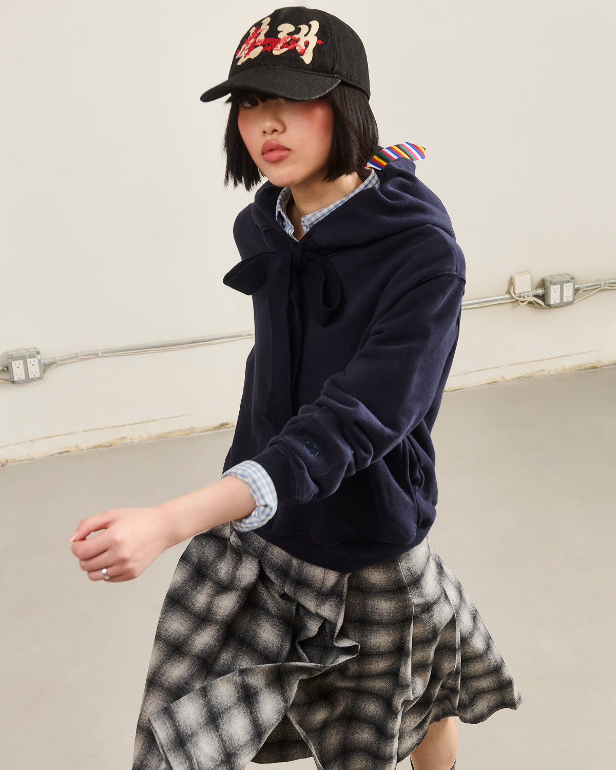 A person wears a black cap, Sundae School NAVY KNIT TIE HOODIE, plaid shirt, and checkered skirt while standing indoors against a plain wall with visible electrical outlets and cords.