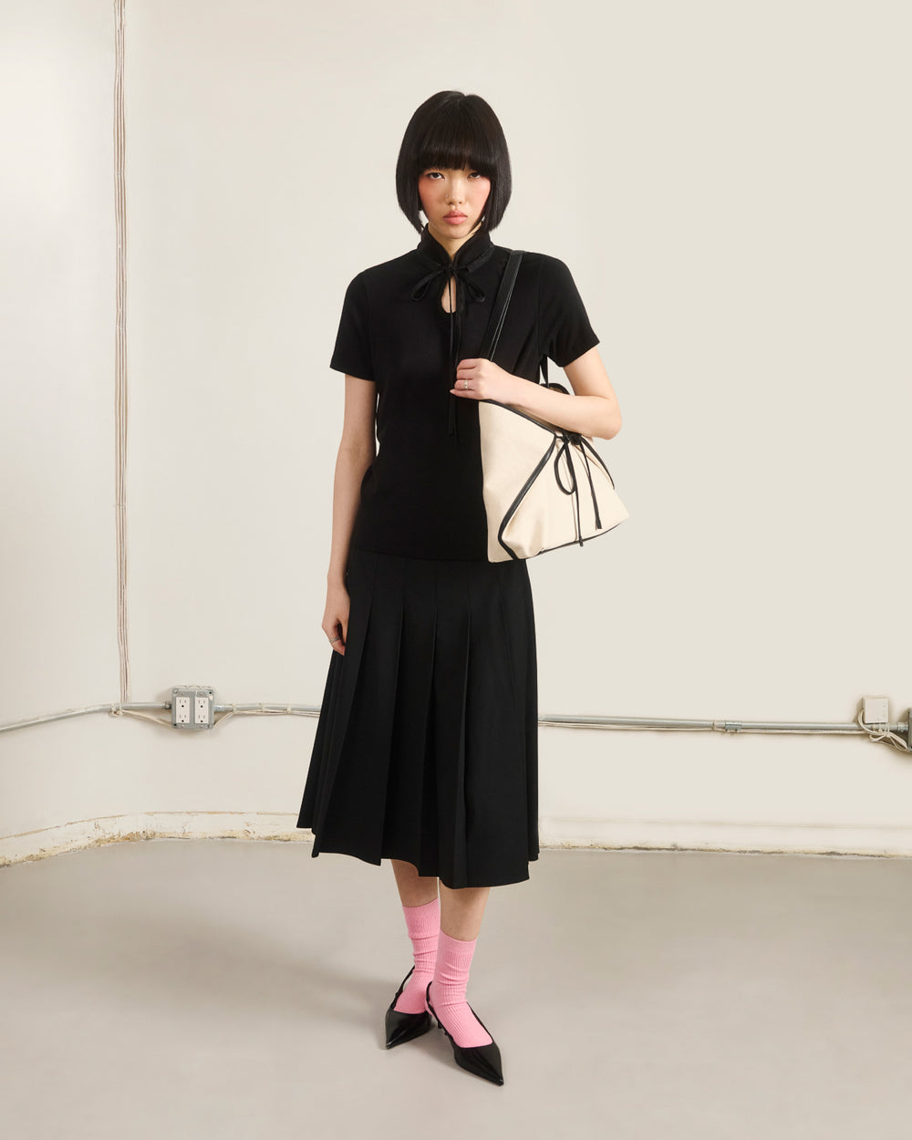 A woman with a short bob wears the Sundae School BLACK RIBBON TIE POLO, a black pleated skirt, pink socks, black pointed shoes, and carries a large cream-colored handbag with black trim in a minimalist room.
