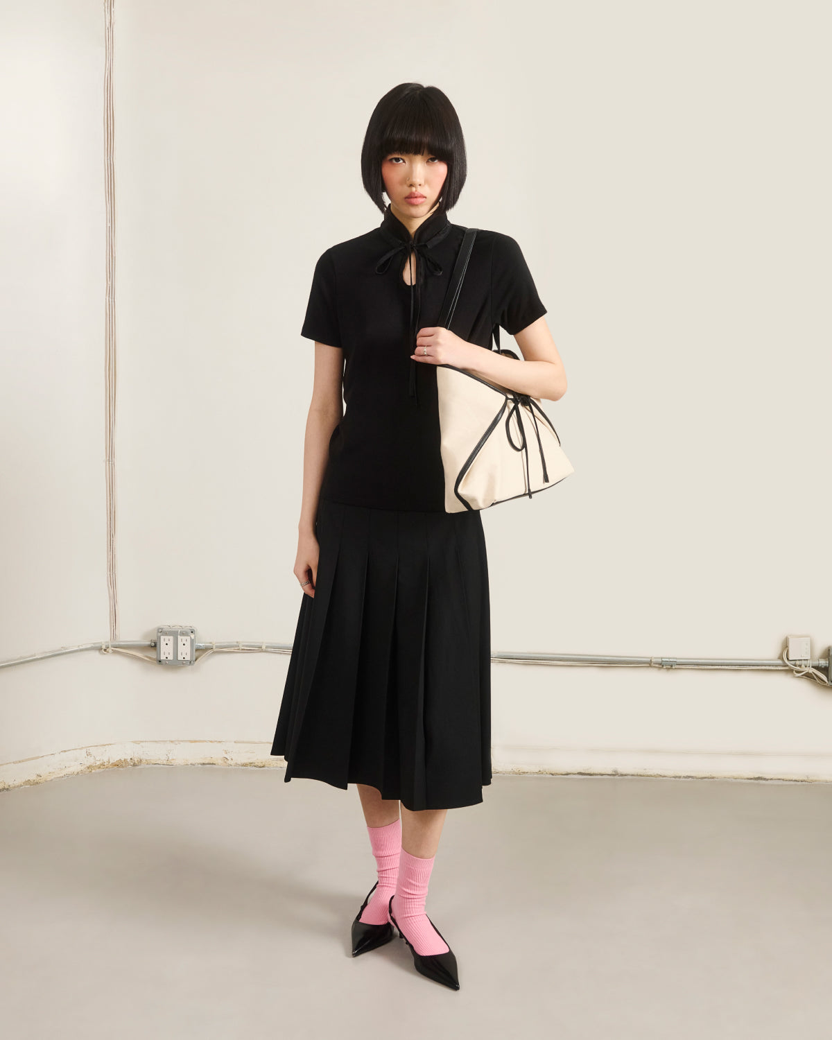 A woman with a short bob wears the Sundae School BLACK RIBBON TIE POLO, a black pleated skirt, pink socks, black pointed shoes, and carries a large cream-colored handbag with black trim in a minimalist room.
