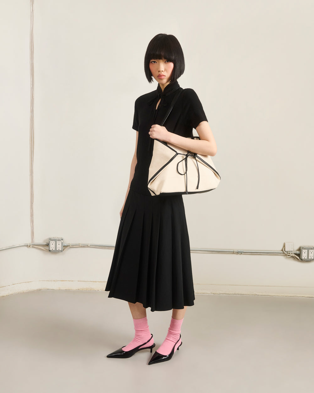 A woman with a short black bob haircut wears the BLACK RIBBON TIE POLO by Sundae School as a dress, paired with pink socks, black pointed shoes, and holds a cream handbag with black trim. She stands against a plain light-colored wall.