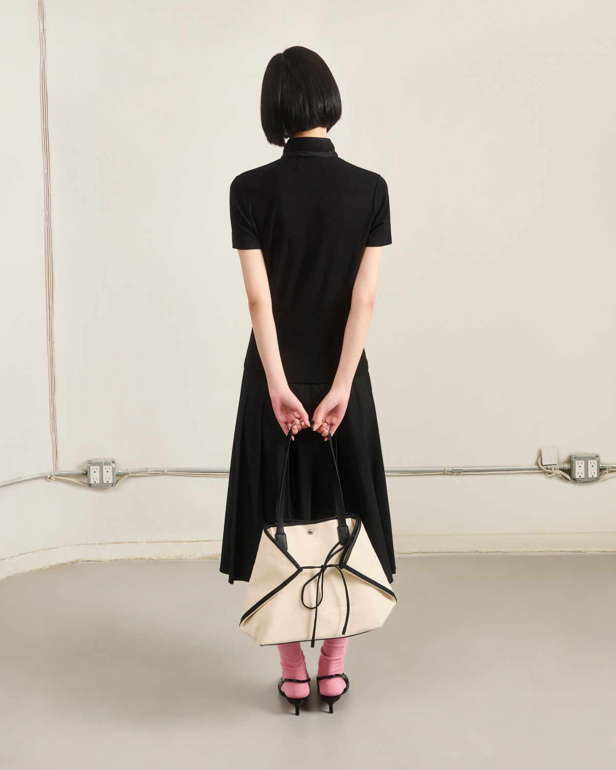 A woman with short black hair wears Sundae School's BLACK RIBBON TIE POLO as a dress, paired with pink socks and heels. Facing away from the camera in a minimalist room, she holds a large cream handbag with black straps behind her back.