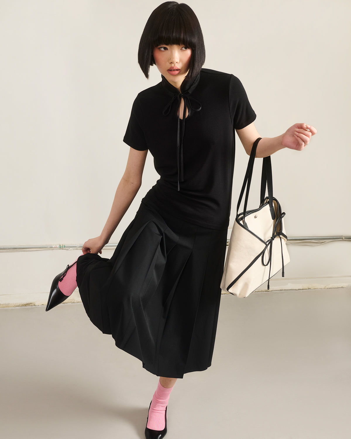 A woman with a bob haircut models Sundae School's BLACK RIBBON TIE POLO, styled with a pleated black skirt, black heels and pink socks, holding a beige tote bag. She poses in a minimal studio with one foot raised, gazing to the side.