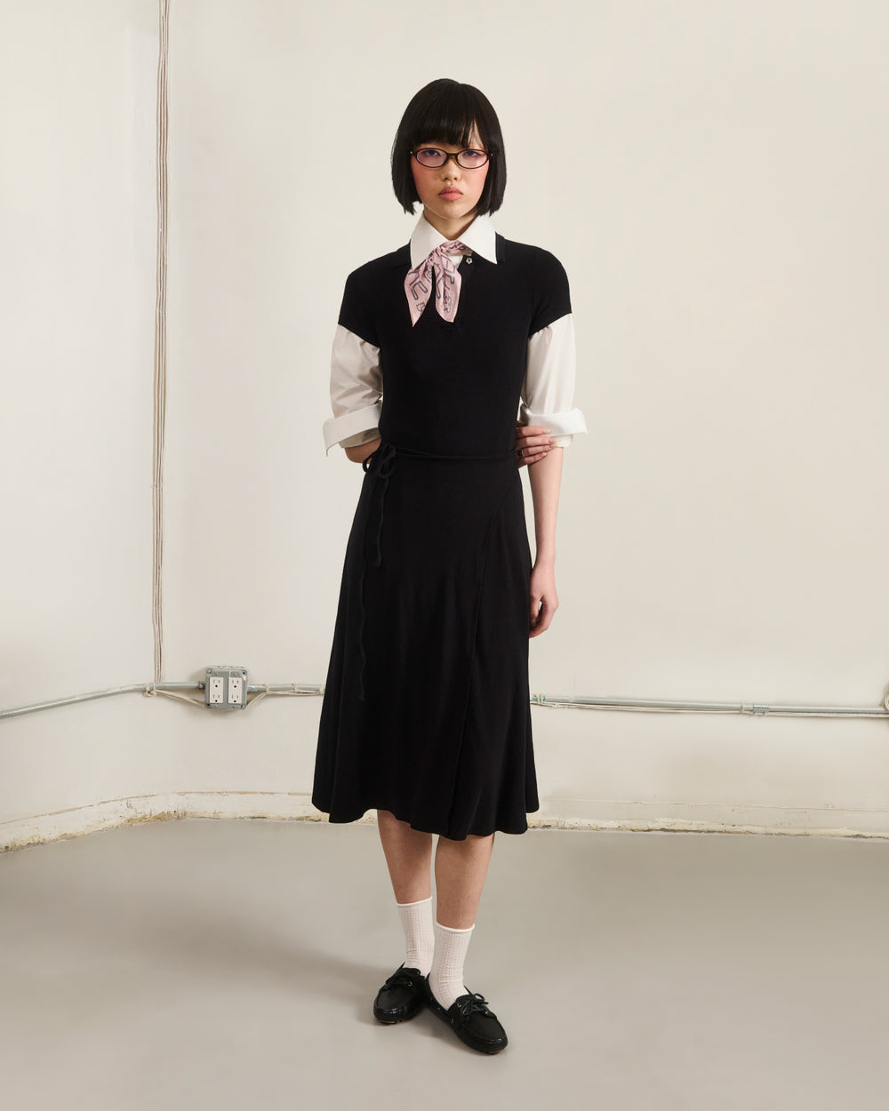 A person with short dark hair and glasses stands against a plain wall in a minimal room, wearing Sundae School’s BLACK CINDY POLO DRESS styled with a white shirt, black sleeveless sweater, pink bow, white socks, and black shoes.