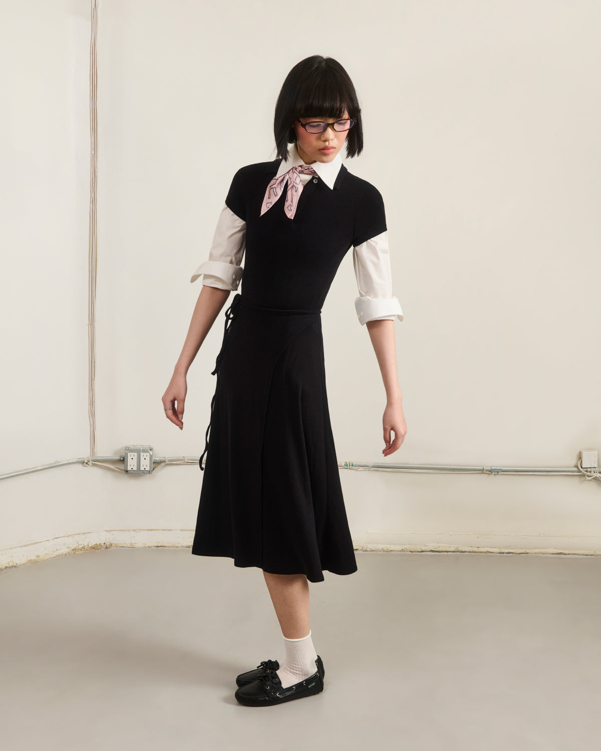 A person with short dark hair, glasses, and a pink scarf wears the Sundae School BLACK CINDY POLO DRESS layered over a white rolled-sleeve shirt, paired with white socks and black shoes, standing in a minimalist room with light walls and visible cords.