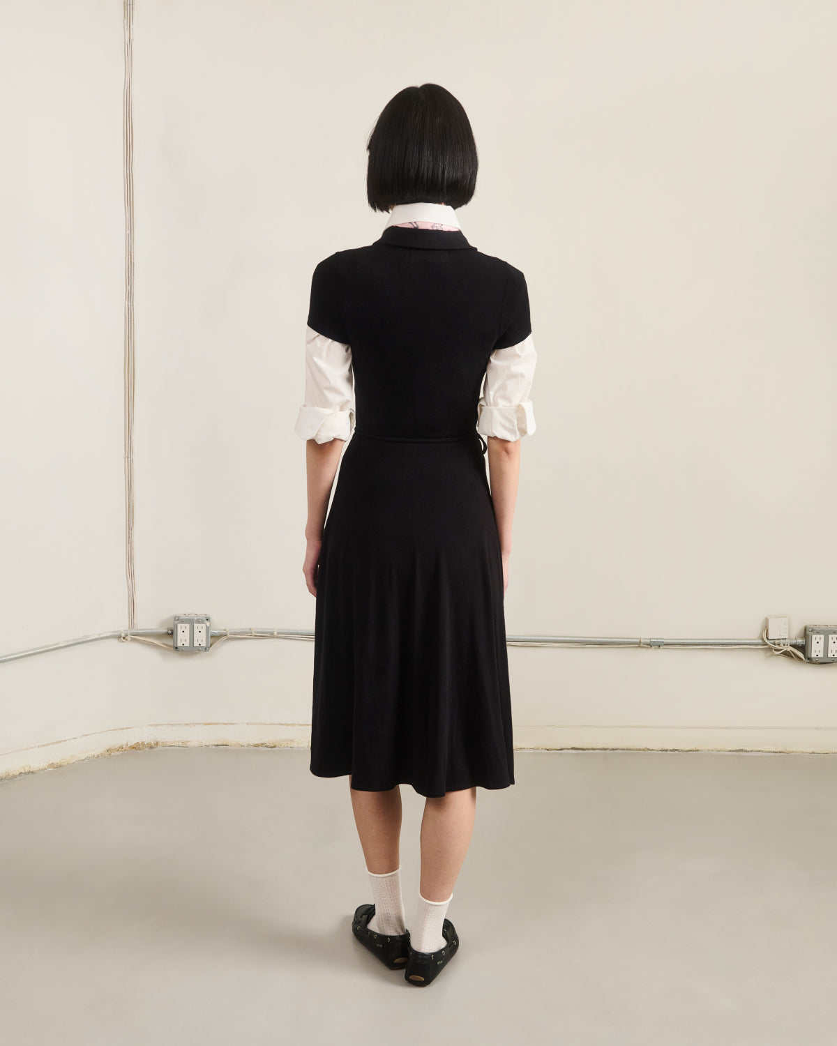 A person with short black hair faces a plain wall, wearing the BLACK CINDY POLO DRESS by Sundae School over a white shirt, paired with white socks and black shoes. Electrical outlets and wires line the base of the wall.