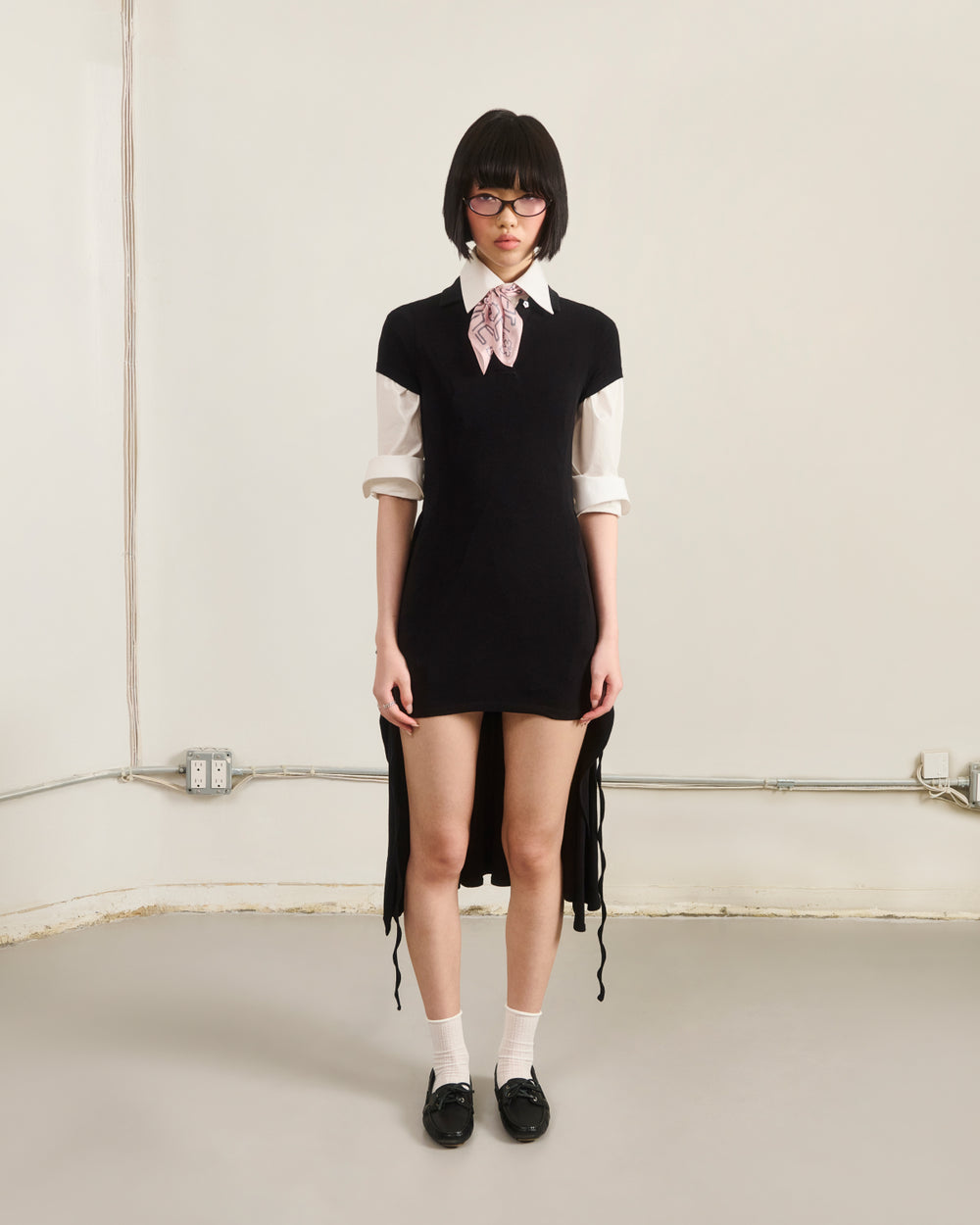 A person with short dark hair, glasses, and a serious expression stands indoors against a plain, light-colored wall, wearing Sundae School’s BLACK CINDY POLO DRESS layered over a white shirt with a pink bow, paired with white socks and black shoes.