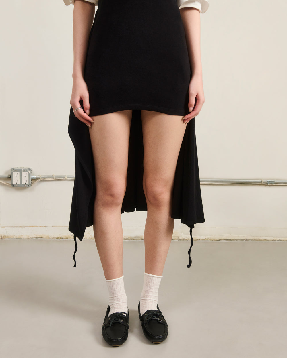 A person stands indoors wearing the BLACK CINDY POLO DRESS by Sundae School—a chic summer wool dress in black with a short front and longer back hem. They pair it with white socks and black loafers, their hands resting by their sides, with only their body from shoulders down visible.