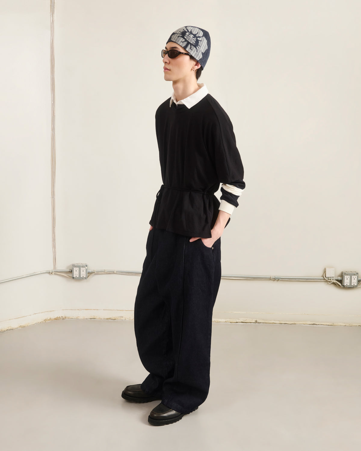 Indoors, a person wears Sundae School’s BLACK BAERAE T-SHIRT over a white collared shirt, with sunglasses, a patterned headscarf, dark loose pants, and black shoes. The plain background shows electrical outlets and pipes.