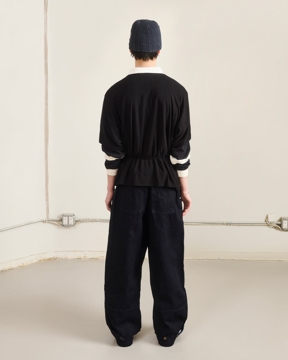A person stands indoors facing a beige wall, wearing a black beanie, loose black pants, and the Sundae School BLACK BAERAE T-SHIRT over a white shirt; electrical outlets and exposed wiring are visible on the wall.