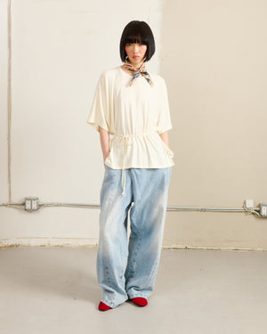 A person with a short black bob wears the Sundae School IVORY BAERAE T-SHIRT belted at the waist, paired with light wide-leg jeans, a patterned neck scarf, and red shoes, standing indoors against a plain beige wall with exposed outlets and cables.