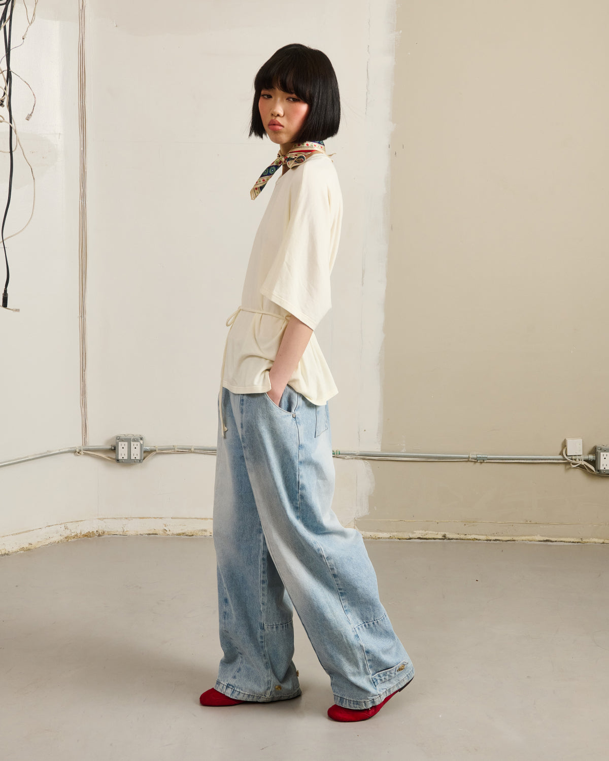 A person with a short black bob stands sideways in a minimal room, wearing the Sundae School IVORY BAERAE T-SHIRT, light blue baggy jeans, red socks, and a patterned scarf around their neck, hands in their pockets.