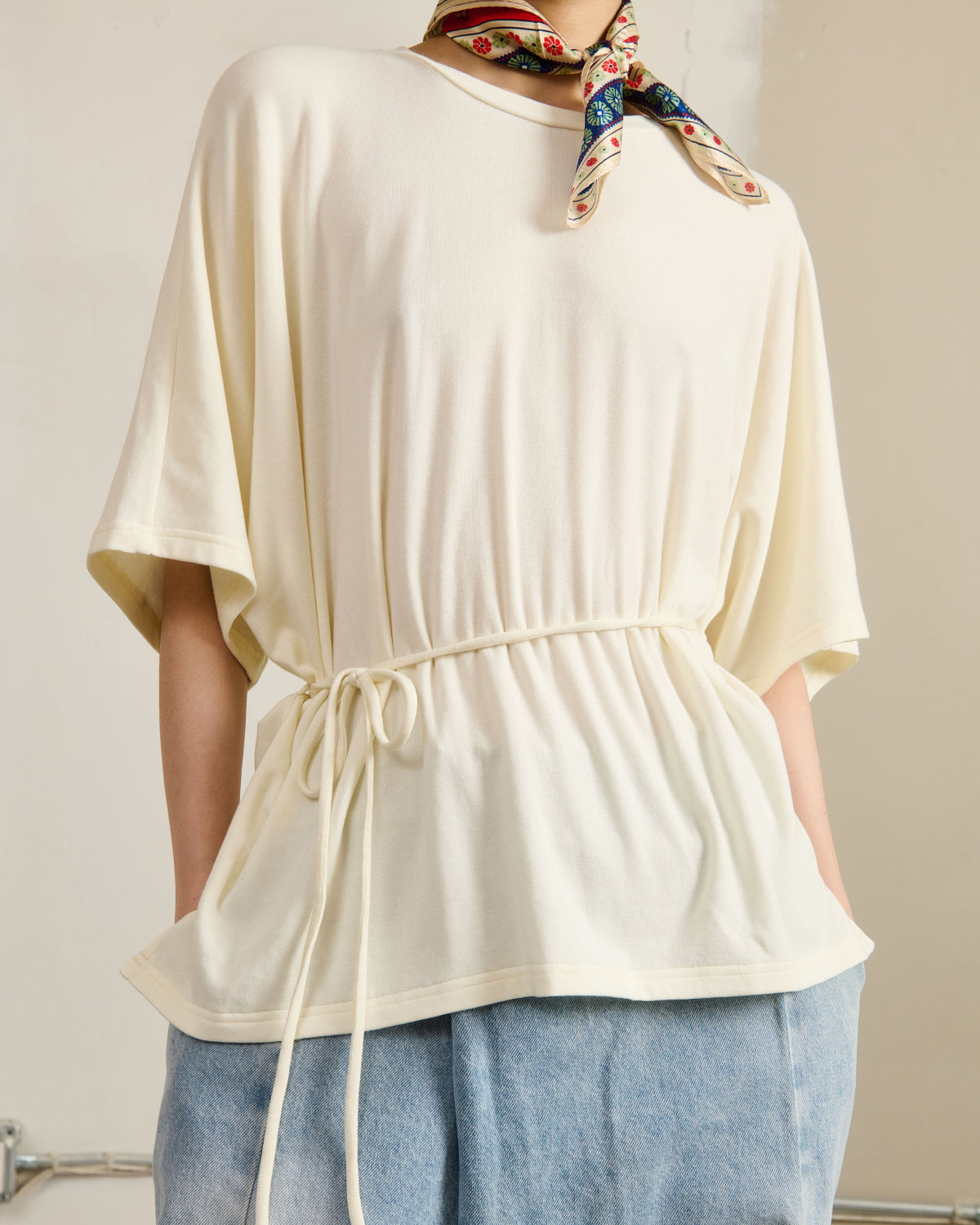 A person wears the Sundae School IVORY BAERAE T-SHIRT with a drawstring waist, light blue jeans, and a colorful patterned scarf tied at the neck, set against a neutral background.