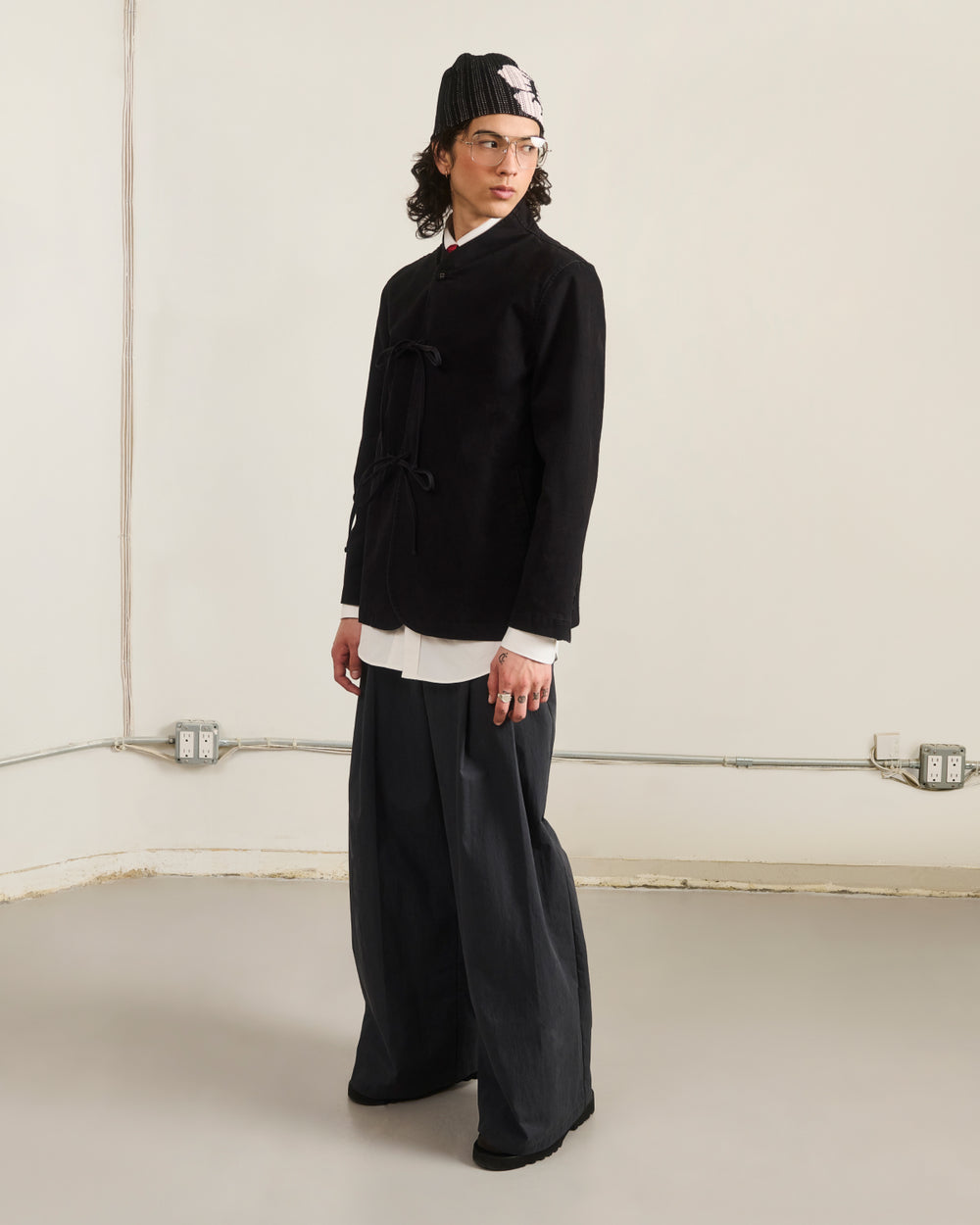 In a minimalist room, a person wears Sundae School’s BLACK JEOGORI TIE JACKET over a white shirt with wide-leg pants, black shoes, a black knit hat featuring a white design, and clear glasses, looking slightly to the side.
