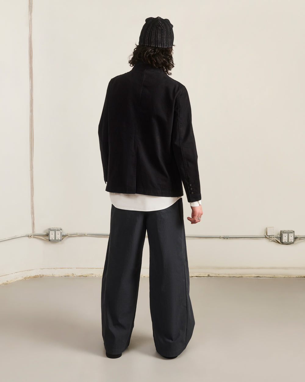 A person stands indoors facing away, wearing Sundae School’s BLACK JEOGORI TIE JACKET over a white shirt, with a black beanie and wide dark pants. Wall outlets and cables are visible along the lower wall.
