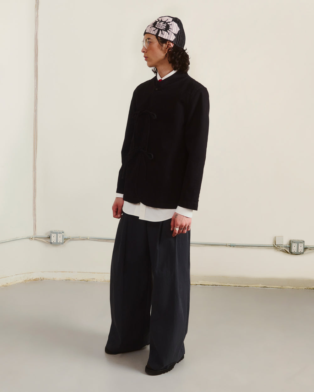 A person wears Sundae School's BLACK JEOGORI TIE JACKET over a white shirt, with loose dark pants, black shoes, and a patterned headscarf, standing indoors against a plain wall while looking to the side.