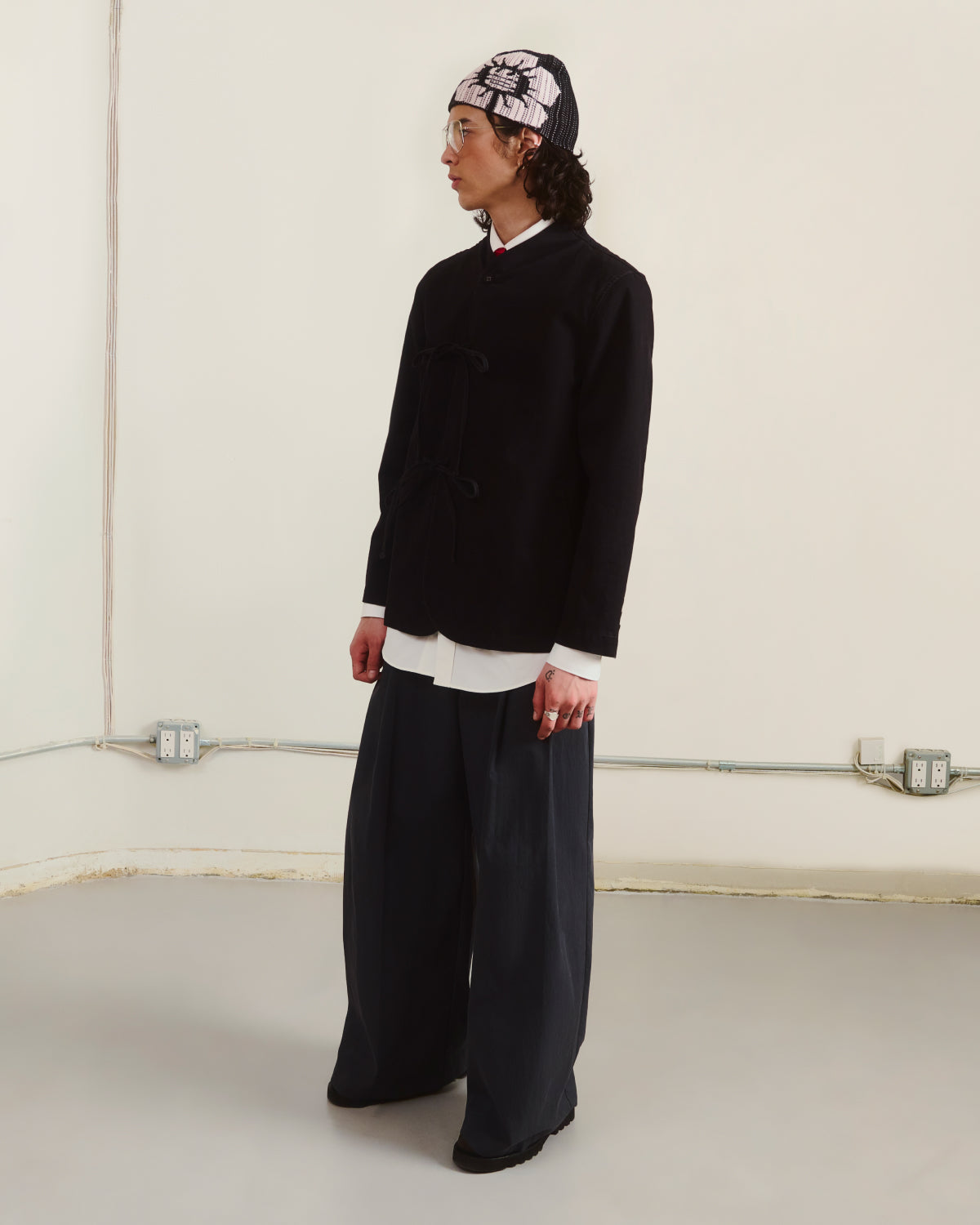 A person wears Sundae School's BLACK JEOGORI TIE JACKET over a white shirt, with loose dark pants, black shoes, and a patterned headscarf, standing indoors against a plain wall while looking to the side.