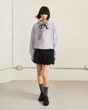 A person with a short black bob stands indoors against a plain wall, wearing the Sundae School HEATHER GREY KNIT TIE HOODIE, paired with a black pleated skirt, gray socks, and black shoes.