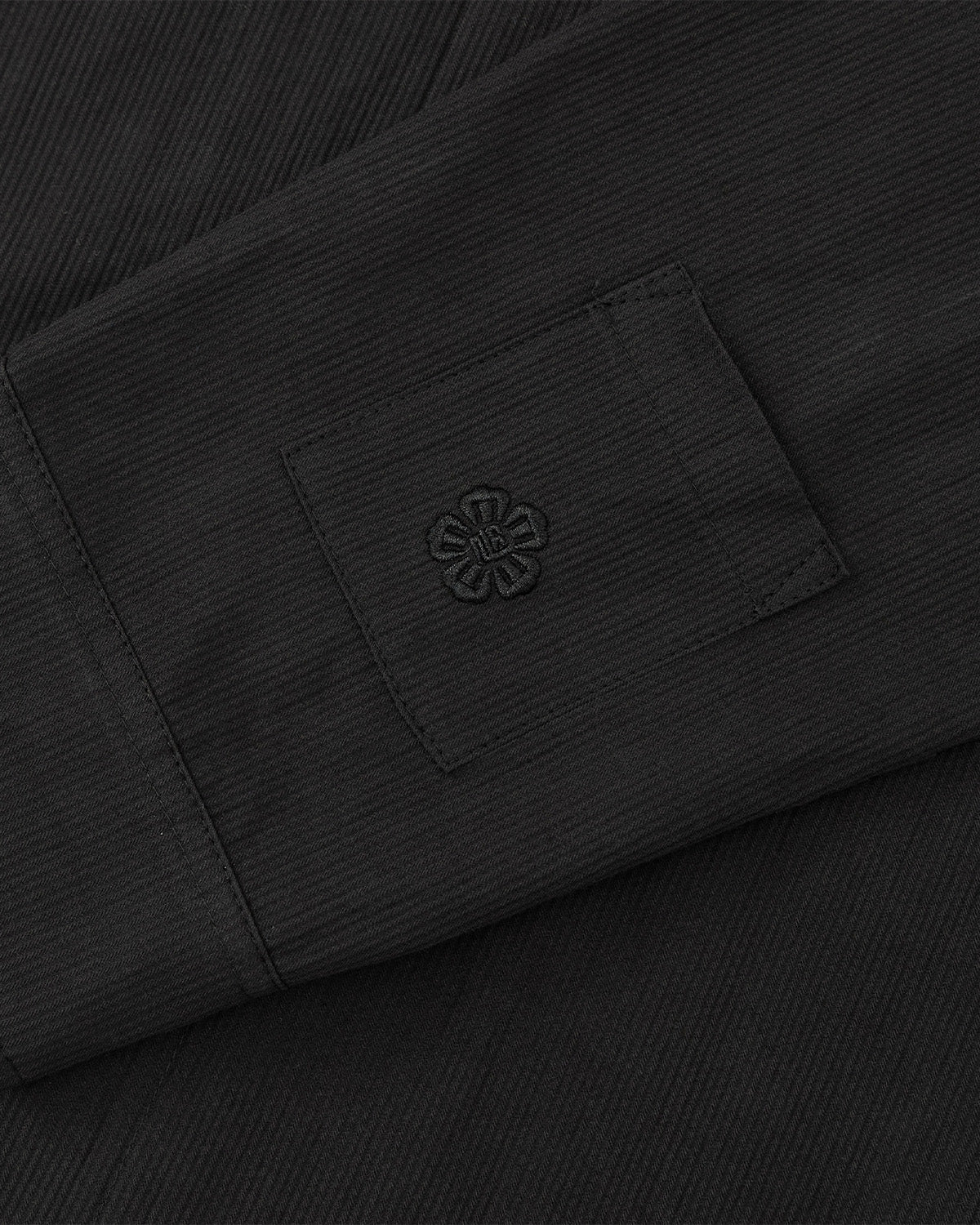 A close-up of the BLACK CHINA COLLAR KNOT SHACKET by Sundae School, showing its black textured fabric with a small rectangular pocket featuring a black embroidered flower-like design.