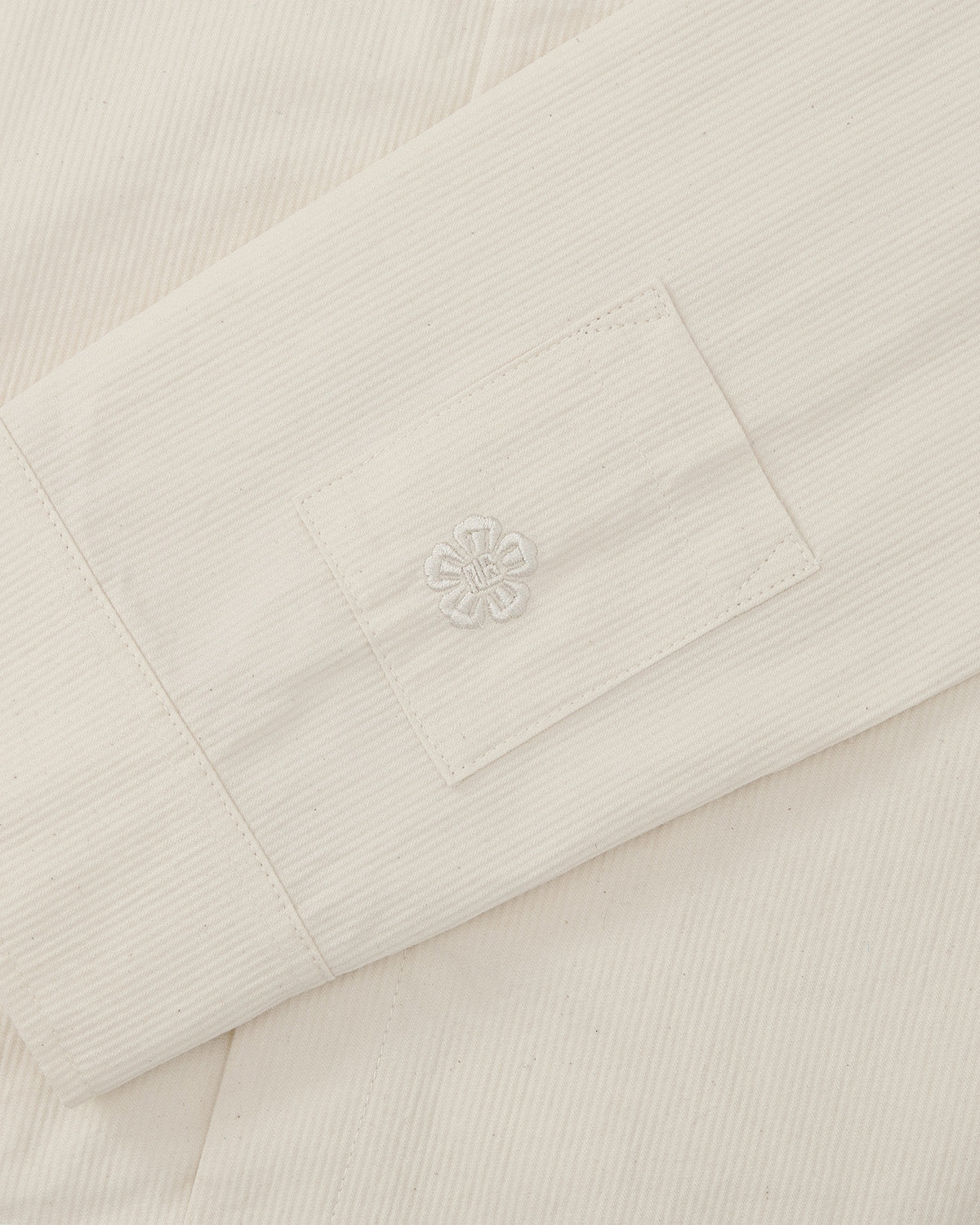 A close-up of the Sundae School IVORY CHINA COLLAR KNOT SHACKET shows its cream corded cotton twill sleeve with a rectangular patch, embroidered flower, and subtle vertical textured lines.