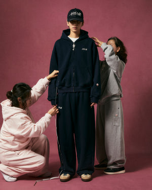 A person wearing the Sundae School NAVY WRAP SWEATBAJI and a cap poses against a pink backdrop while two others in light pink and gray wide-leg sweatpants adjust clothing and measure.