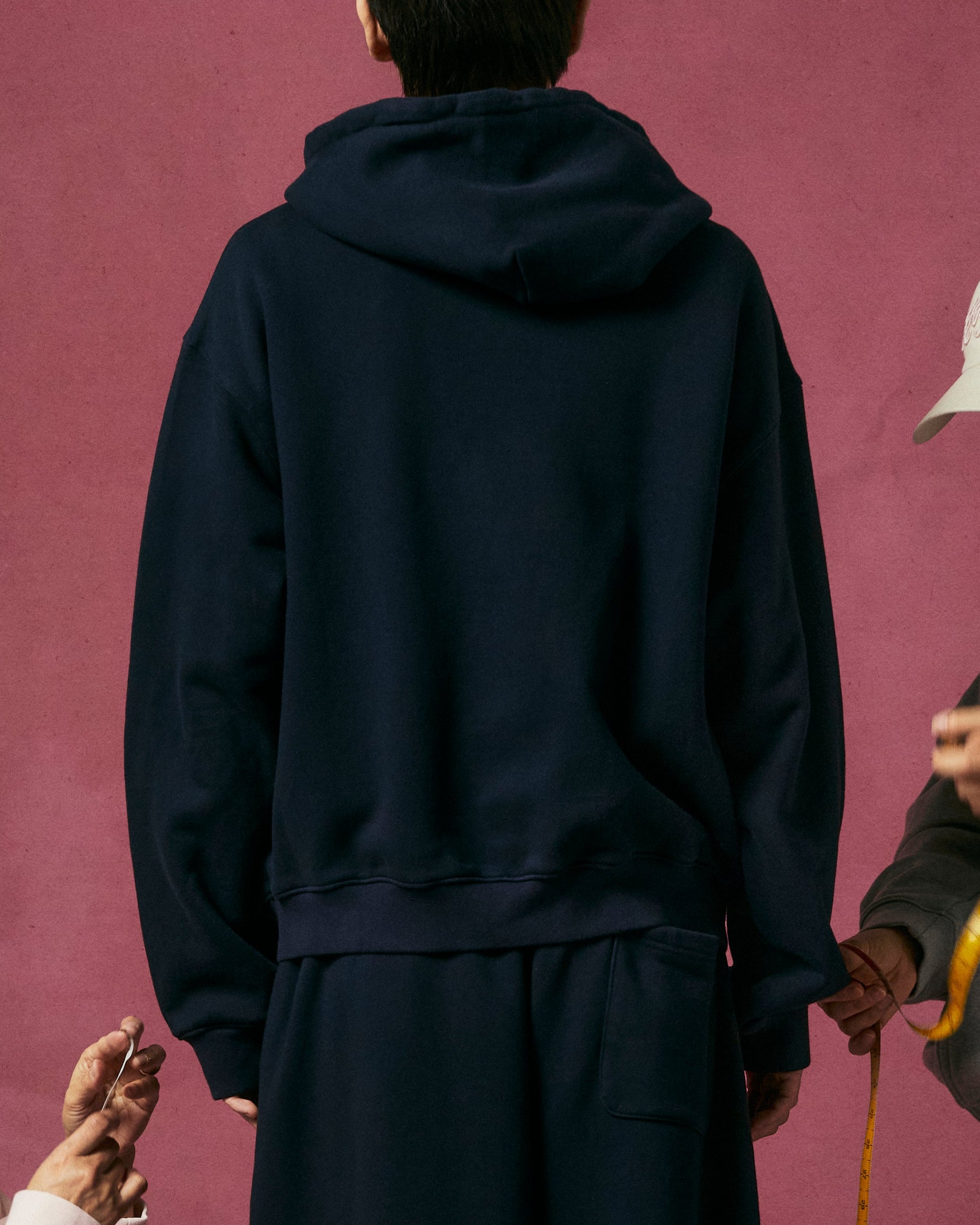 Three people in Sundae School casuals pose before a pink backdrop. One, dressed in the NAVY WRAP SWEATBAJI, stands facing away, while two others in light pink and gray use measuring tapes to measure the person at center.